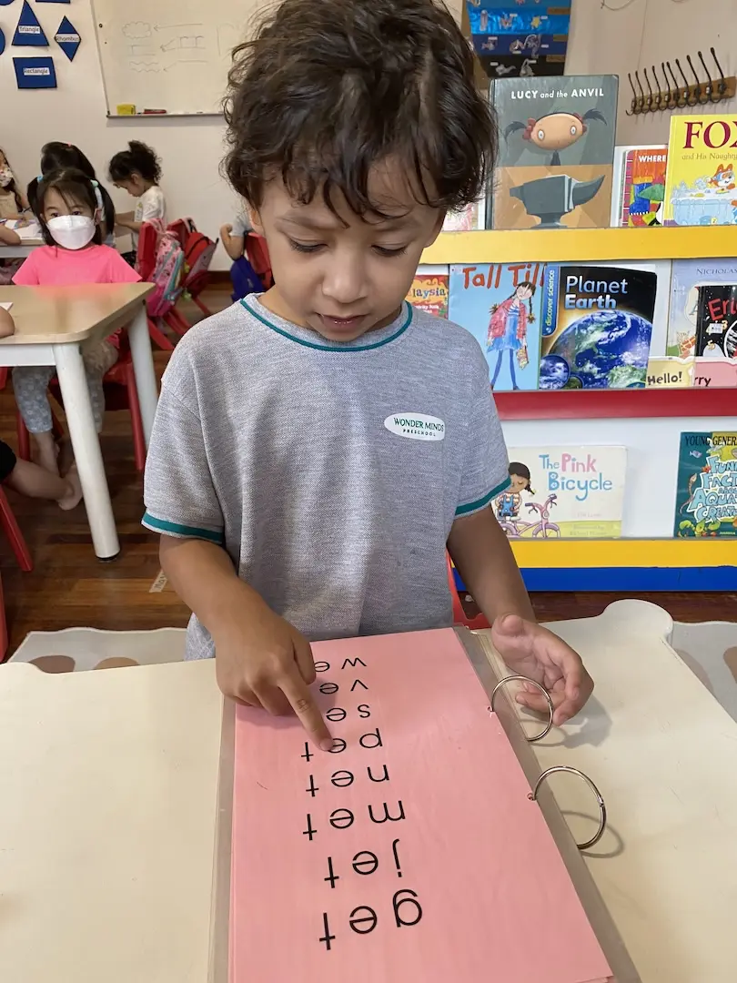 A boy pointing to letters on a pink card in front of a bookshelf